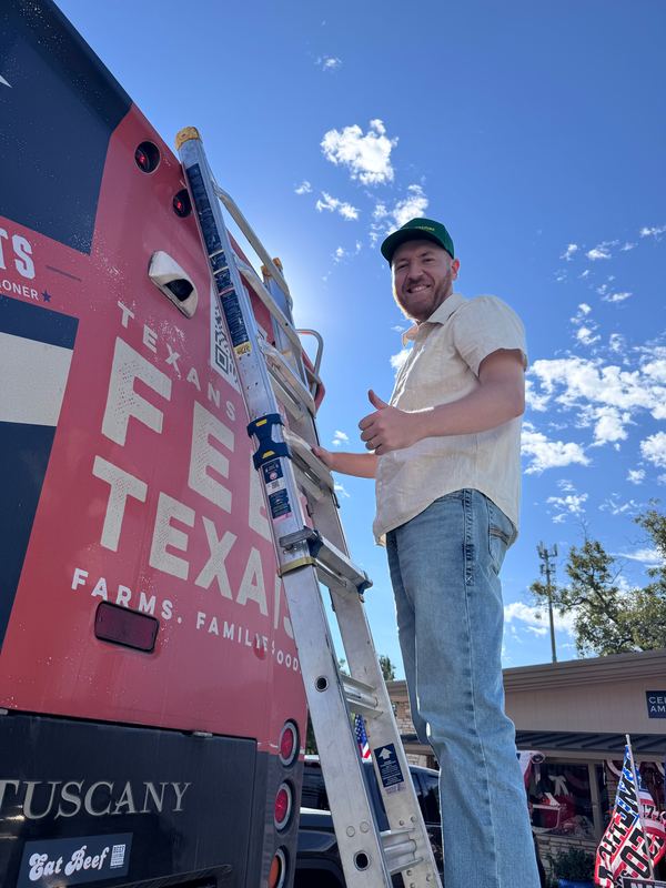 Austin Jensen giving a thumbs up from a ladder beside the red Texas Feeds Texas bus